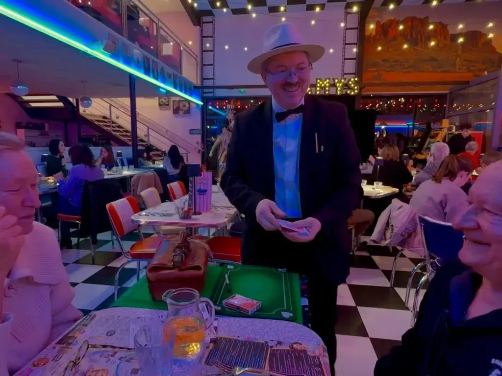 Sylvio, en veste sombre, debout dans un restaurant de style américain, tient quelques cartes devant des convives attablés