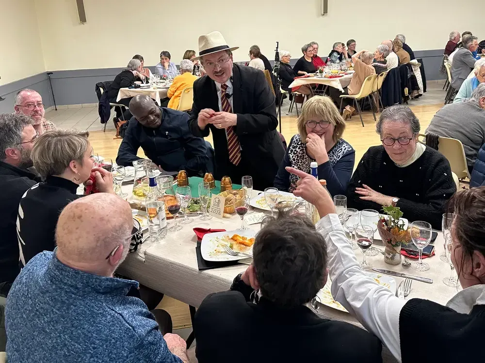 Sylvio, chapeau panama et cravate, montre un tour à une table de convives lors d’un repas associatif en salle des fêtes