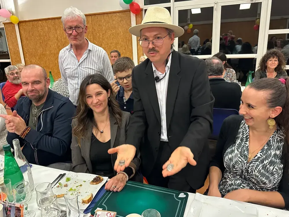 Sylvio, micro-casque sur la joue, tient deux pièces au-dessus d’une table de convives souriants
