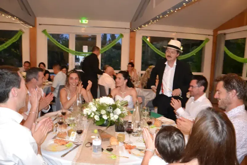 Des convives de mariage applaudissent autour de tables fleuries, tandis que Sylvio, chapeau panama, se tient debout dans la salle