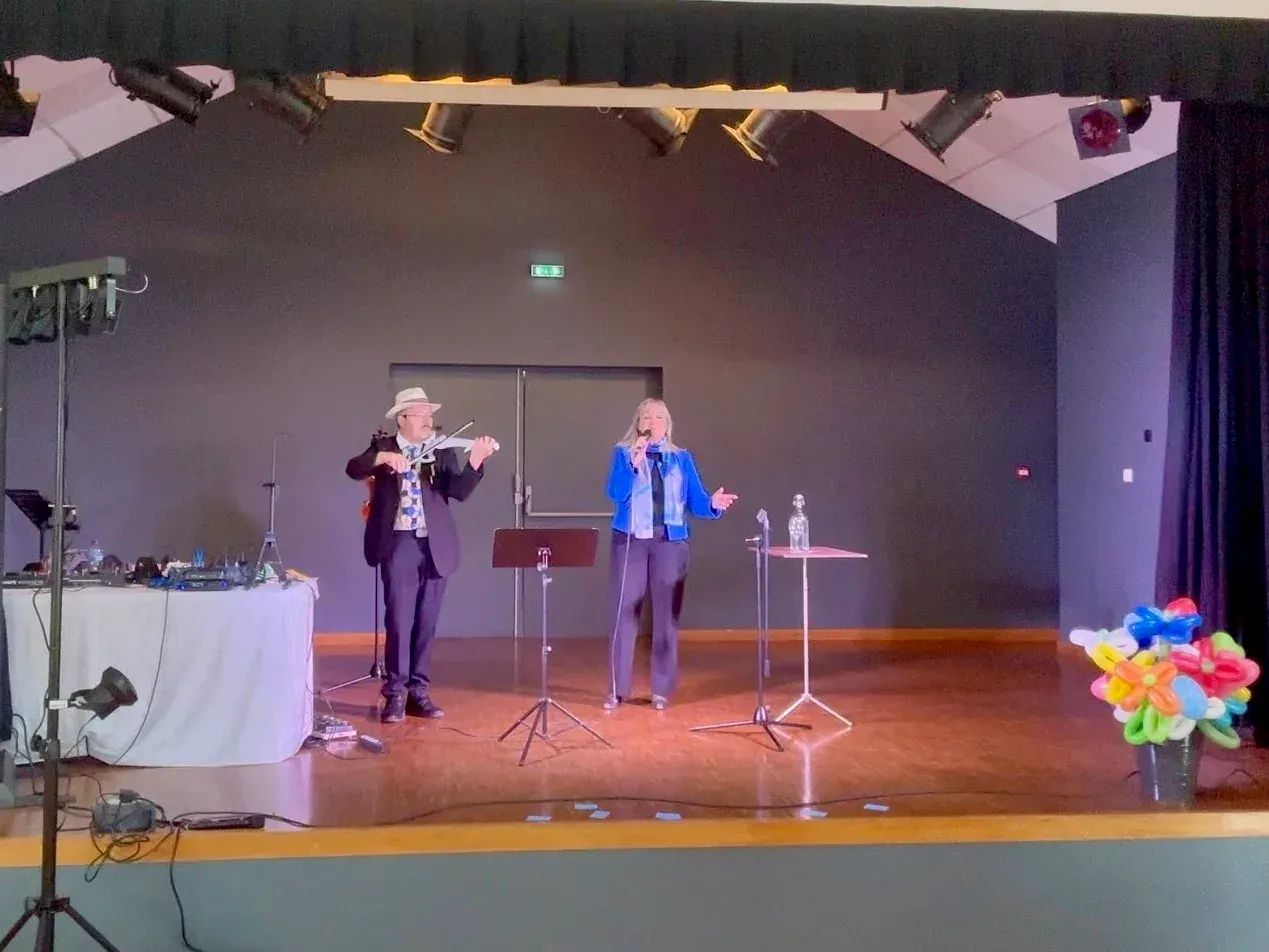 Sylvio au violon et Nathalie au micro sur la scène d’une salle, près d’un bouquet de ballons sculptés