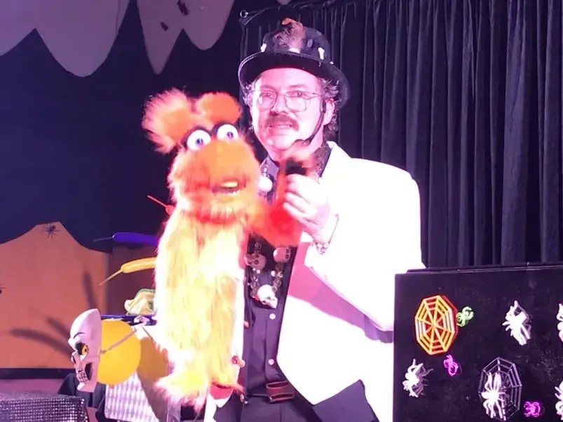 Sylvio en veste blanche et chapeau noir, une marionnette orange à la main, dans le décor du spectacle d'Halloween