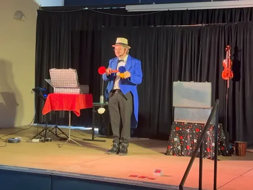 Sylvio, en queue de pie bleue, tient deux pompons rouge et bleu sur scène, entre deux grandes malles décorées et un violon