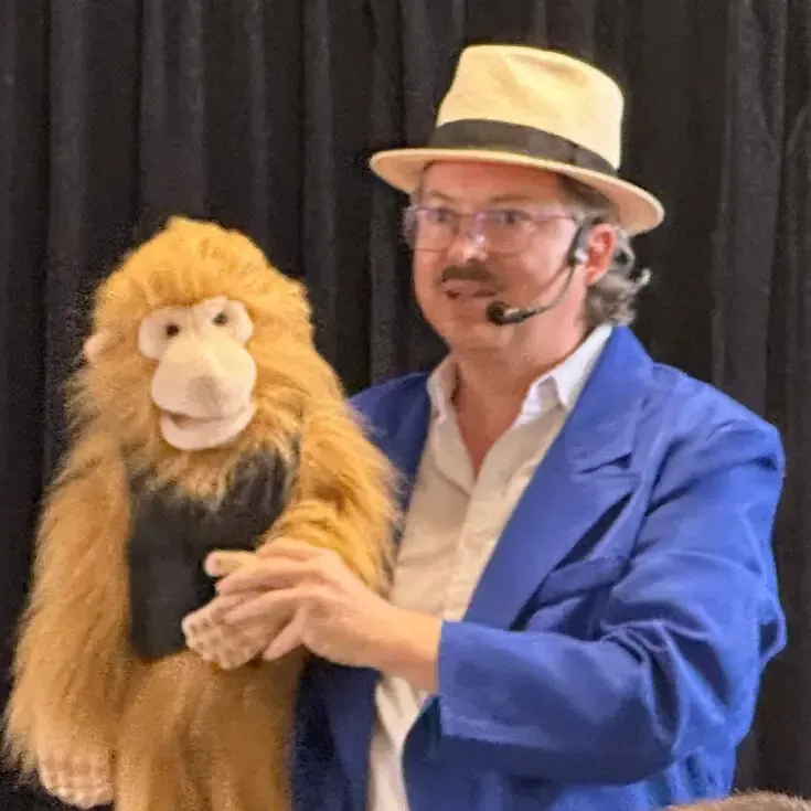 Sylvio, chapeau panama sur la tête et micro-casque, tient dans ses bras une marionnette de singe