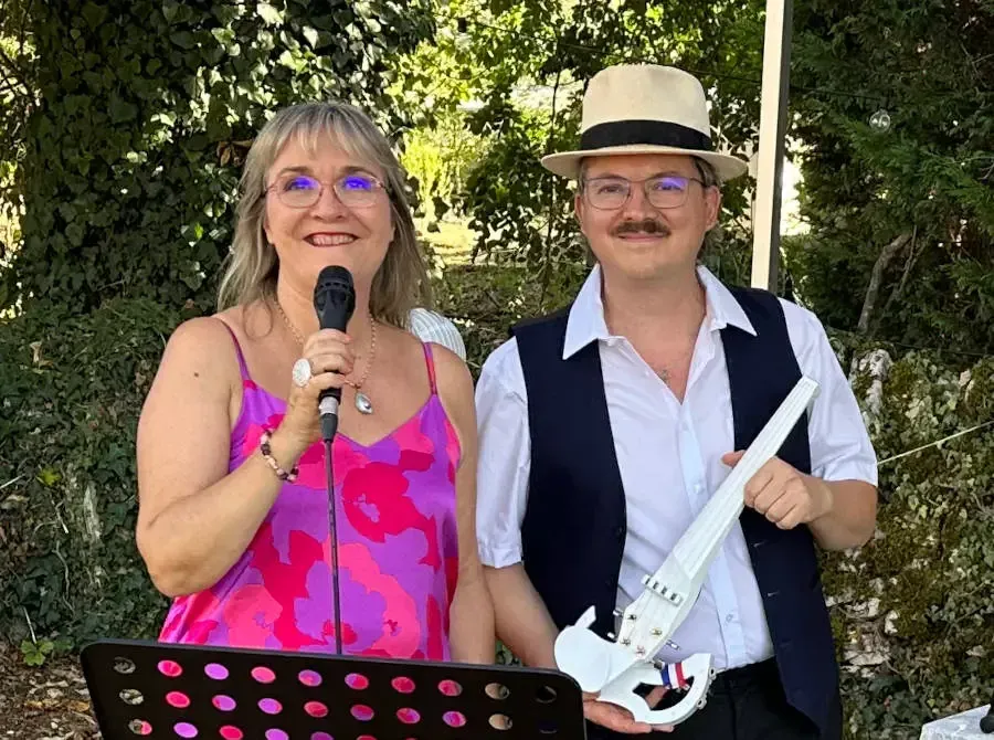 Nathalie, micro à la main, et Sylvio, gilet noir et chapeau panama, tenant un violon blanc, posent dans un jardin