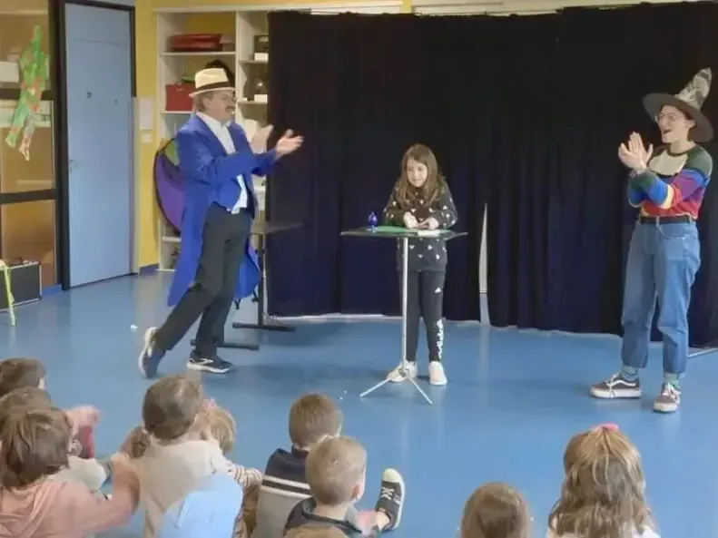 Sur scène, une enfant fait un tour entre Sylvio et une assistante, devant le public d'enfants assis