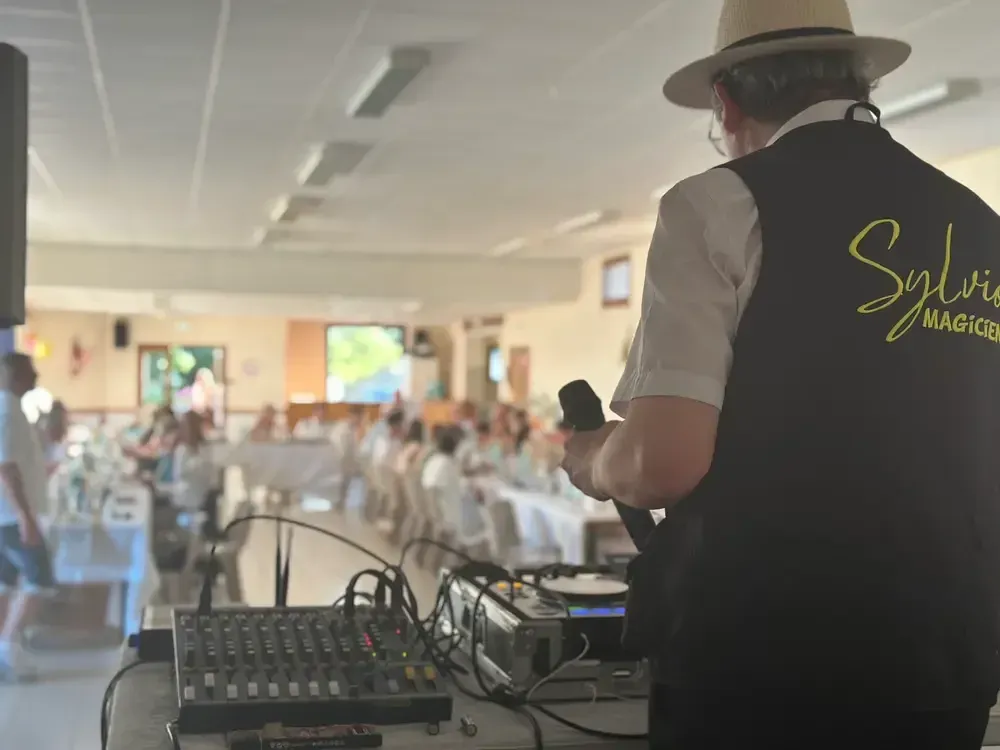 Sylvio, de dos et en gilet noir brodé à son nom, devant une table de mixage, dans une salle de réception dressée pour un repas