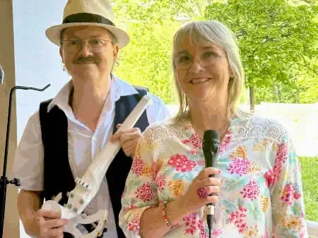 Sylvio, chapeau panama et gilet noir, tient un violon blanc à côté de Nathalie, micro à la main, en chemisier fleuri
