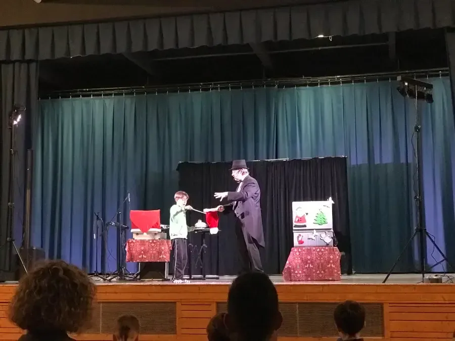 Sylvio, haut-de-forme noir, fait un tour avec un garçon sur la scène d'une salle des fêtes, aux coffres décorés de Noël