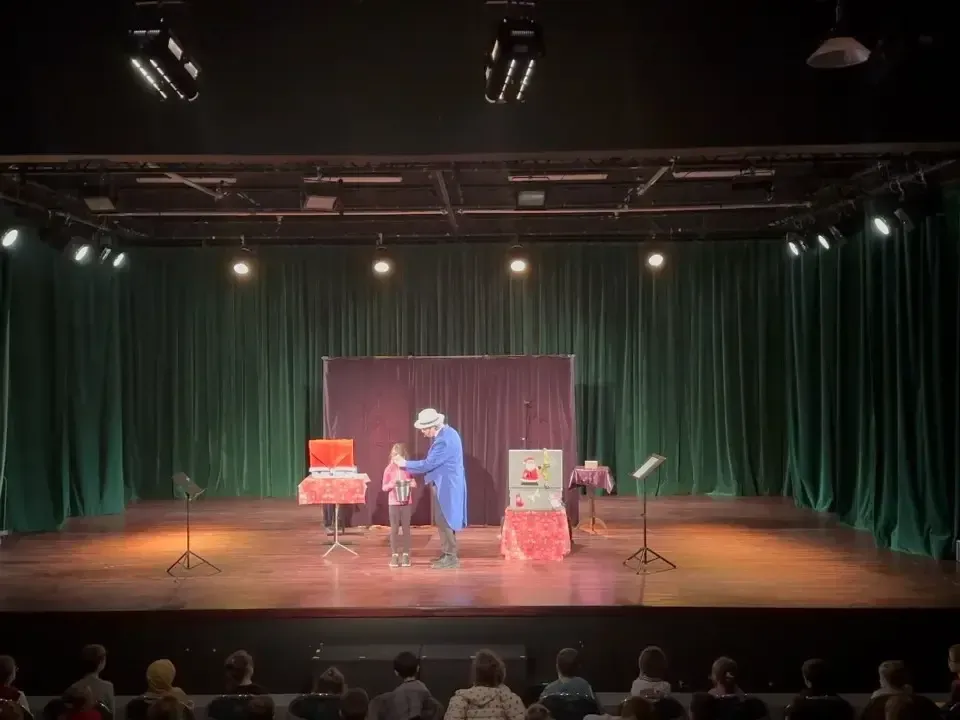 Vue de la salle d’un spectacle de Noël : Sylvio, en veste bleue, sur une grande scène aux rideaux verts avec deux décors de Noël