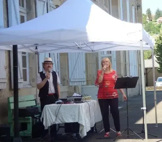 Nathalie chante au micro et Sylvio se tient près d'une table de sonorisation, sous une tente blanche, dans une cour