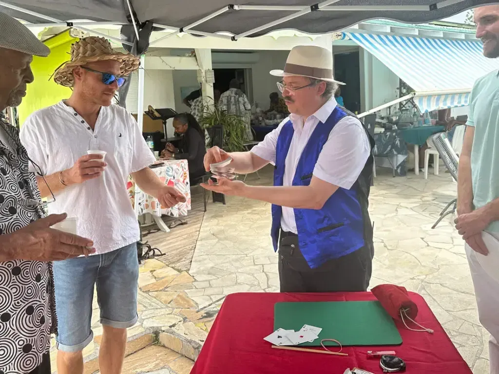 Sylvio, gilet bleu et chapeau panama, fait un tour avec des gobelets devant des convives, sous une terrasse couverte