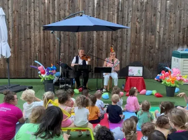 Sylvio au violon et Natymi chantent sous un parasol, devant de jeunes enfants assis sur des tapis, au milieu de ballons