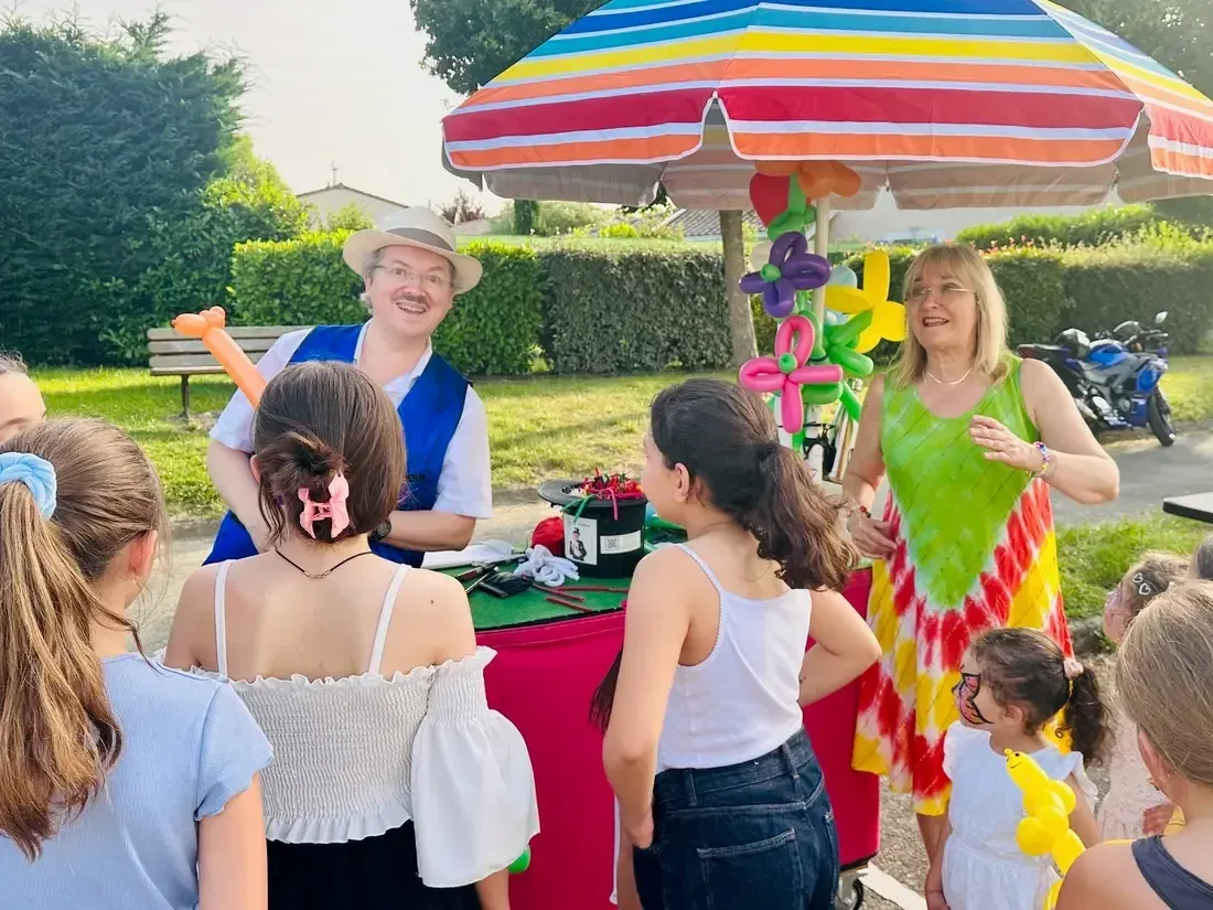 Sylvio et Nathalie en plein air sous un parasol coloré, entourés d’enfants, devant le chariot de magie et de ballons