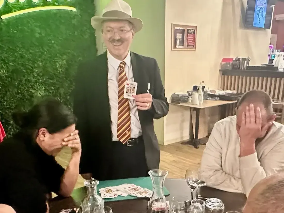 Sylvio, chapeau panama, montre une carte à des clients de restaurant ; l'une d'elles se couvre le visage en riant