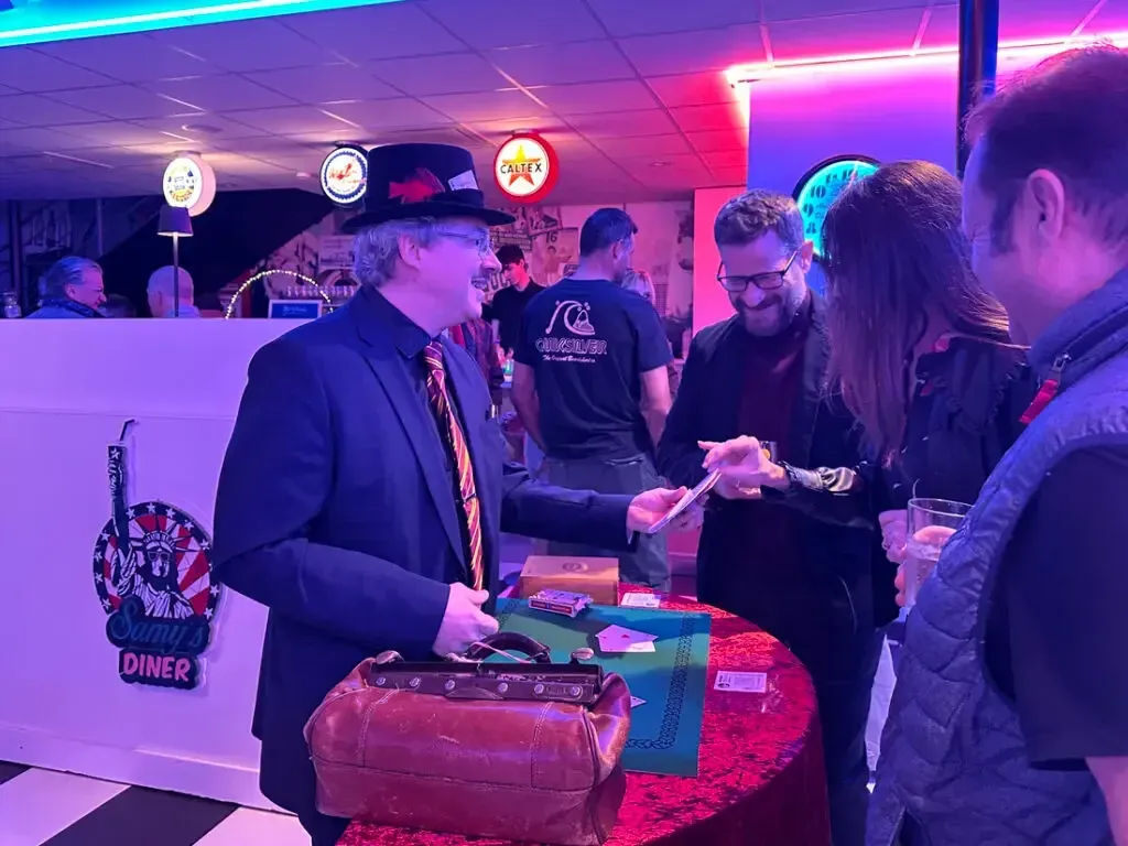 Sylvio, en costume sombre, présente un tour de cartes à des convives dans une salle éclairée de rose et de bleu