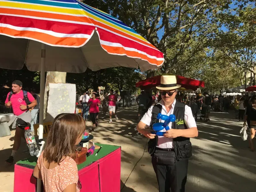 Sylvio, gilet noir et chapeau panama, tient un chien en ballon bleu sous un parasol coloré, une enfant de dos devant lui