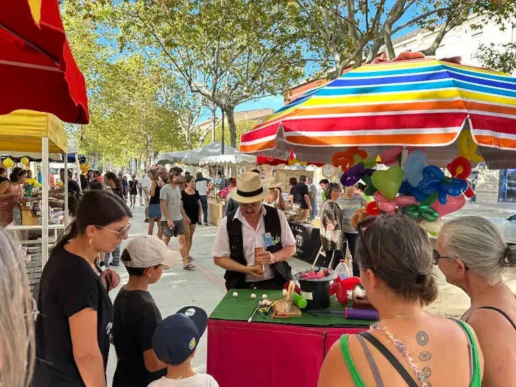 Sylvio à son stand de magie et de ballons, sur un marché ombragé, des familles autour de la table verte