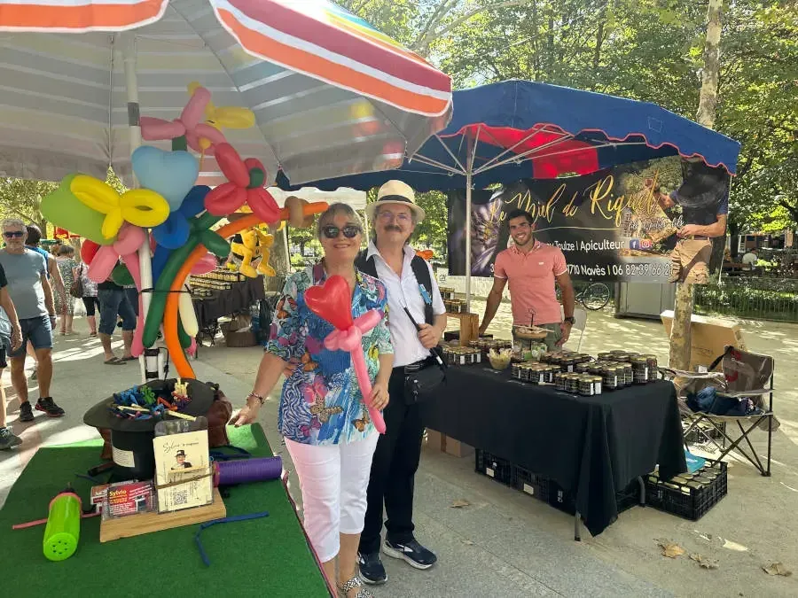 Nathalie et Sylvio posent devant leur stand de ballons et de magie, sous des parasols, sur un marché