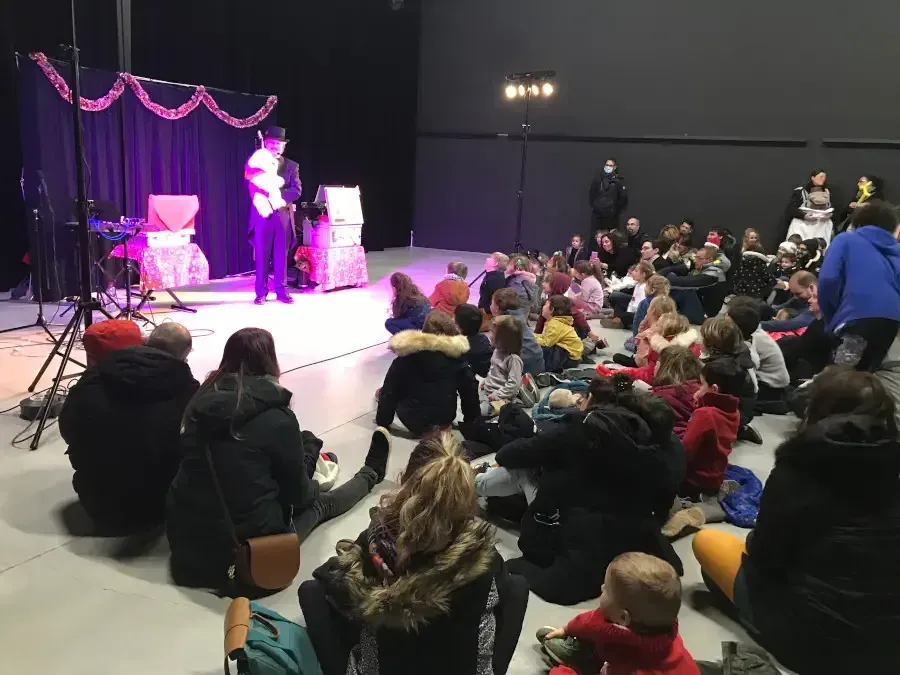Sylvio sur scène sous des guirlandes roses, devant des enfants et des parents assis par terre, vus de dos