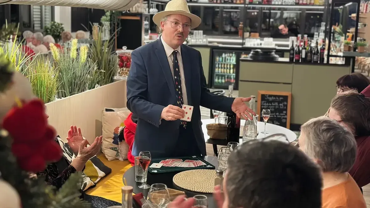 Sylvio, chapeau panama et cravate, montre une carte à des clients de restaurant ; l’une d’elles se couvre le visage en riant