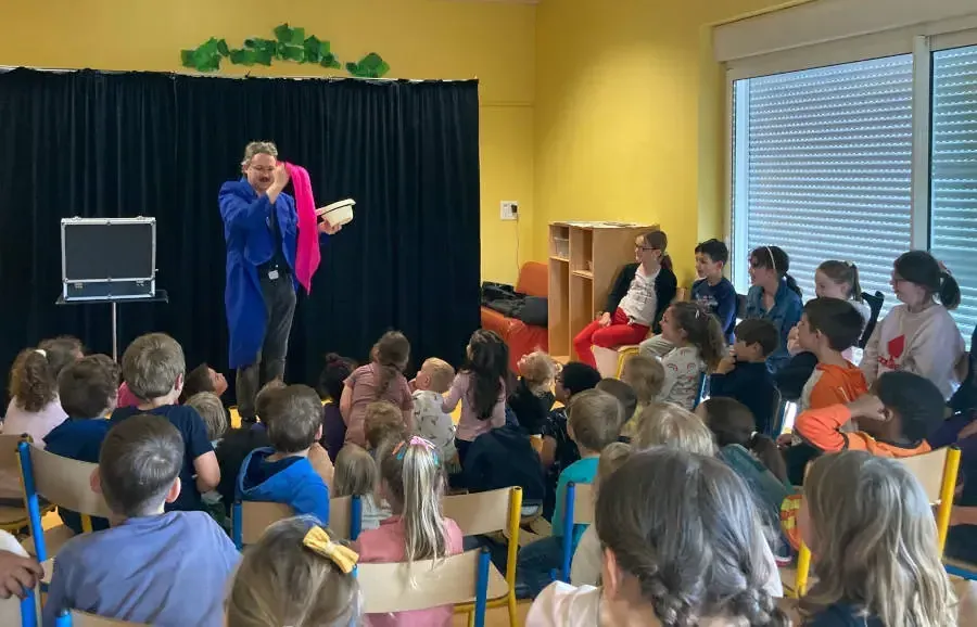 Sylvio, un foulard rose à la main, devant des enfants assis dans une salle d'activités aux murs jaunes