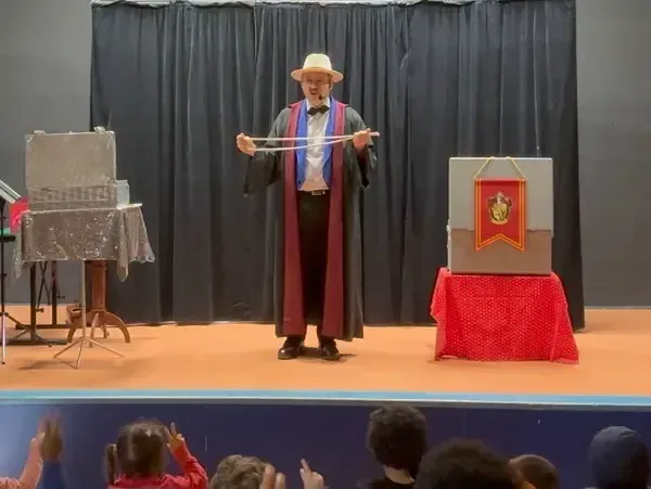 Sylvio, en robe sombre et chapeau panama, tend les bras sur scène entre un coffre gris et un coffre rouge décoré