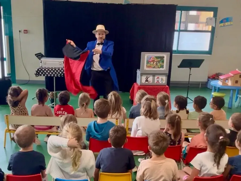 Sylvio fait un tour avec une cape rouge devant des enfants de maternelle assis sur des bancs, vus de dos
