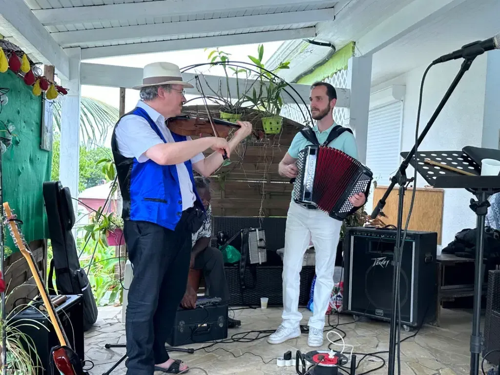 Sylvio au violon et un accordéoniste jouent sous une terrasse couverte, devant un pupitre et un micro